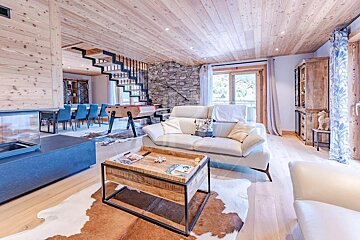 Modern chalet living room featuring wood paneling, stone accent wall, white sofas, foosball table, fireplace, dining area, and open stairs.
