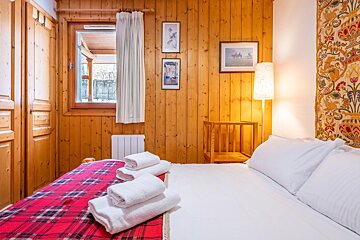 A cozy, wood-paneled bedroom features a bed with white bedding, a red plaid blanket, and fresh towels. A window and art adorn the walls.