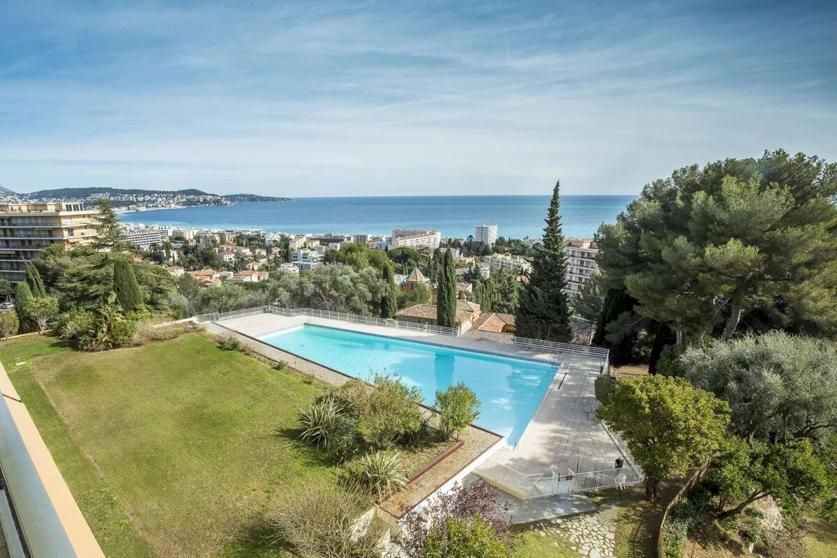 Elevated view of a long swimming pool, green lawn, and coastal city with the blue sea and sky in the background.