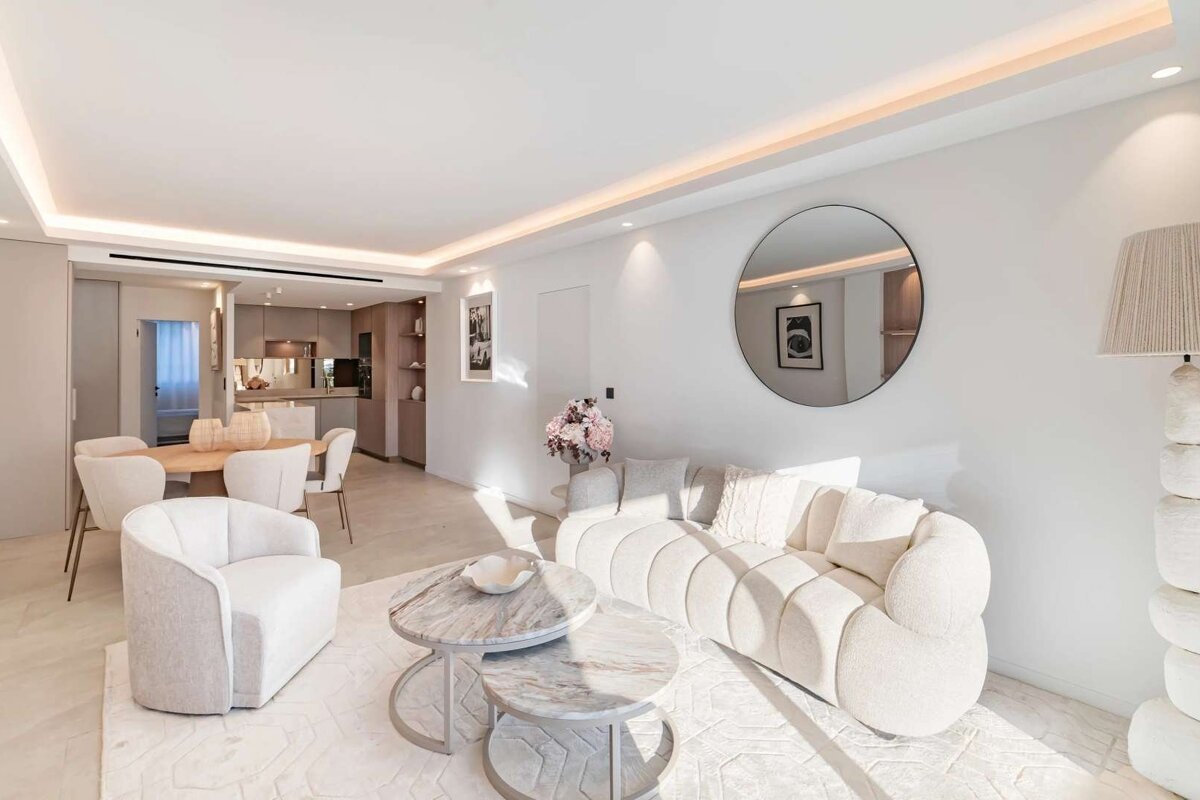 A chic, modern open-plan living and dining area in neutral tones. Features include a curved sofa, marble tables, and subtle lighting.