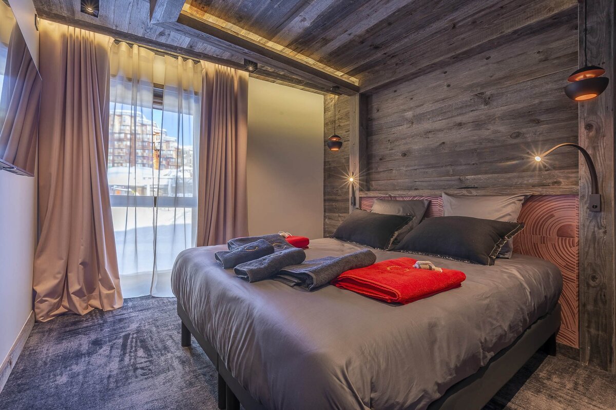 A cozy modern bedroom with dark wood accents, a bed with red towels, and a large window overlooking a snowy landscape.