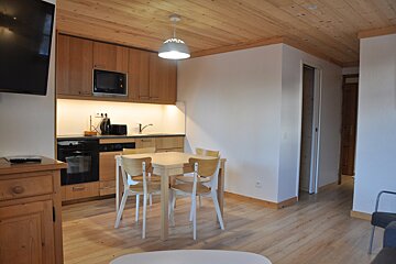 A modern studio apartment featuring a compact wooden kitchen, a dining table with four chairs, light wooden floor and ceiling, and white walls.