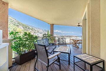 Outdoor balcony with seating & planters, overlooking a scenic coastline, city, and mountains under a blue sky.