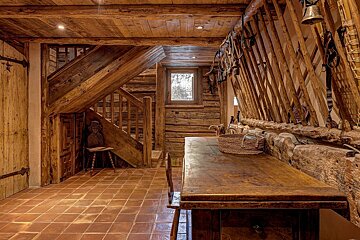 A rustic wooden interior features a prominent staircase, log walls, a large table, and terracotta tiled floor. Bells and straps adorn a unique railing near a small window.