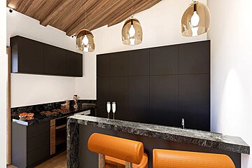 A stylish modern kitchen with black cabinets, dark marble countertops, orange bar stools, and a wooden ceiling. Pendant lights illuminate champagne flutes.