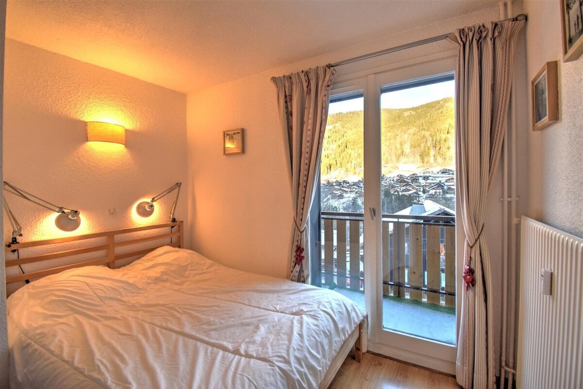 Cozy bedroom with a white bed, warm wall lighting, and balcony doors showing a snowy mountain village view.
