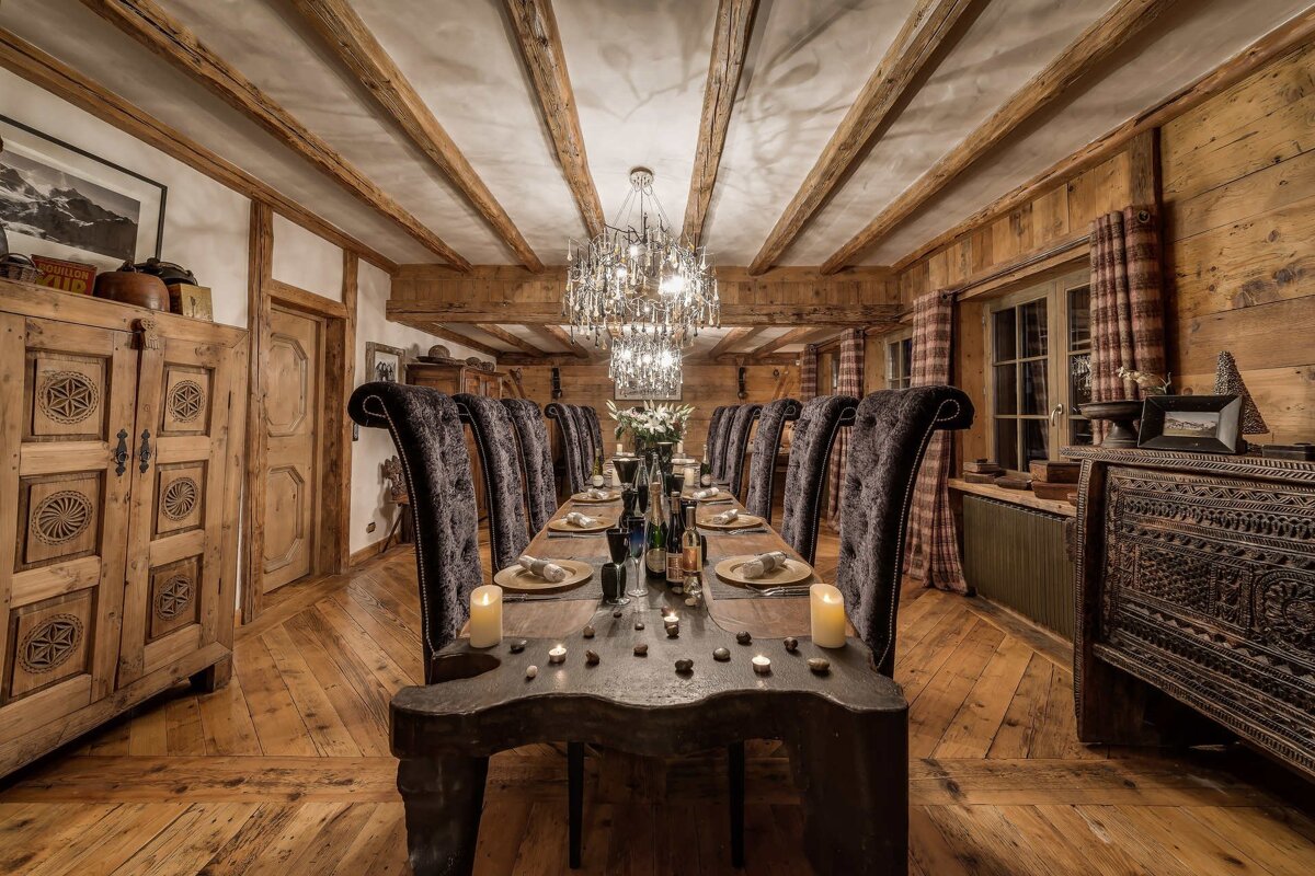 A cozy chalet-style dining room with a long table, high-back velvet chairs, exposed wooden beams, and a grand crystal chandelier.