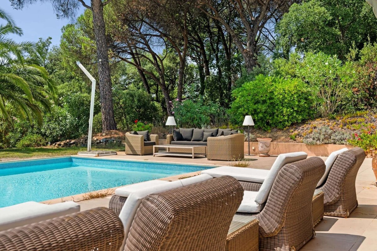 An inviting outdoor space with a sparkling pool, comfortable wicker patio furniture, and sun loungers, set against a backdrop of lush greenery.
