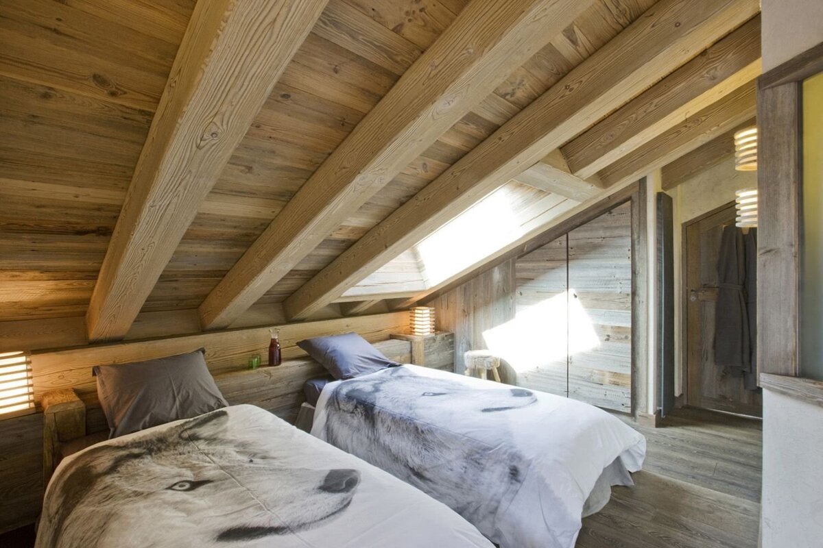 A cozy, rustic attic bedroom featuring warm wooden walls, exposed beams, a skylight, and two beds with unique wolf-print duvet covers.