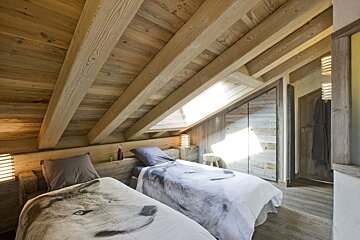 A cozy, rustic attic bedroom featuring warm wooden walls, exposed beams, a skylight, and two beds with unique wolf-print duvet covers.