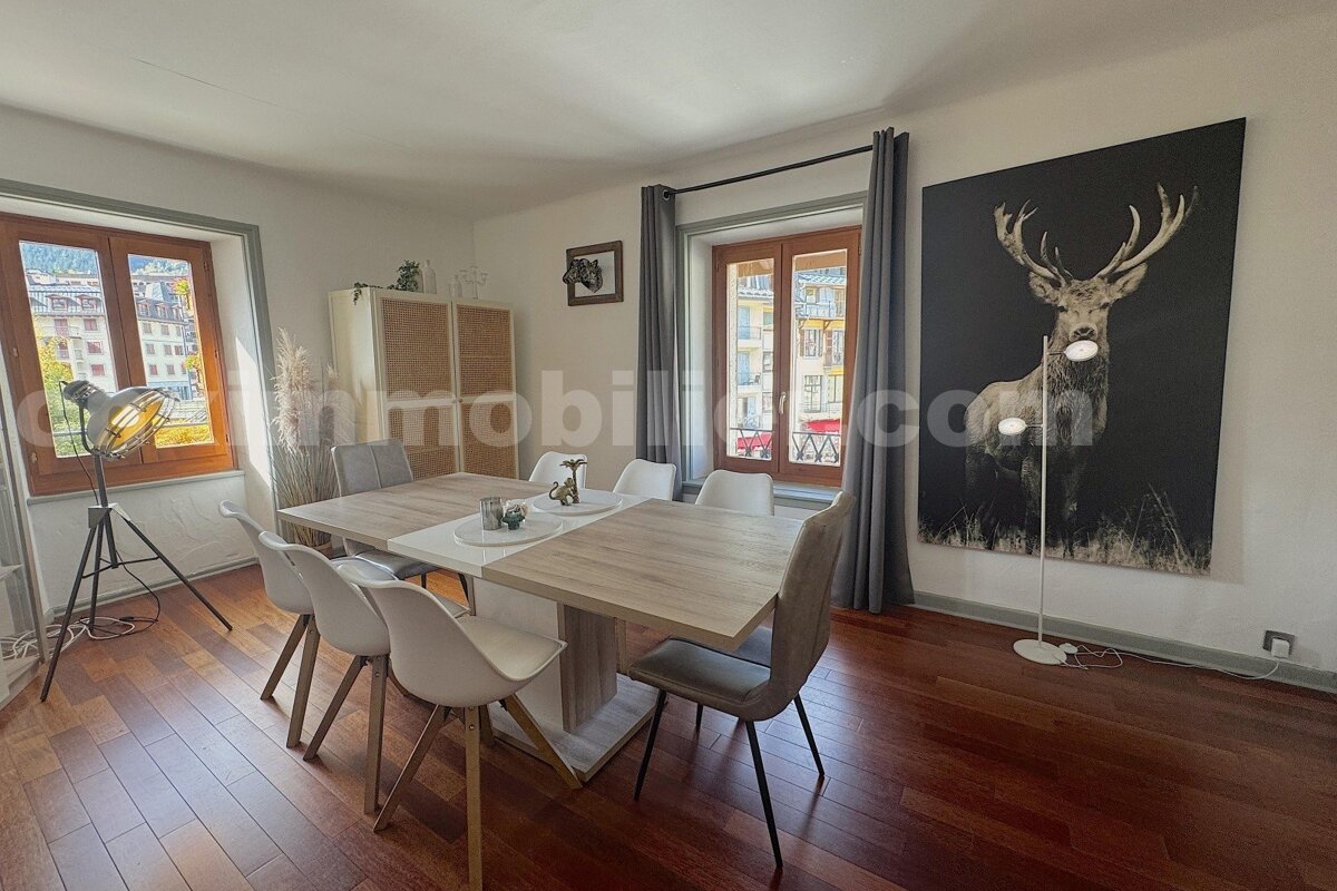 A modern dining room features a wooden dining table with chairs, hardwood floors, two windows, and a large black and white deer artwork on the wall.