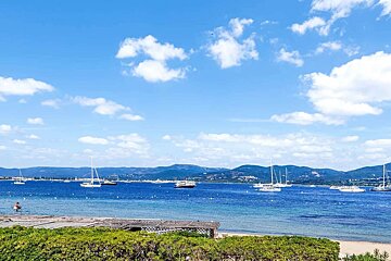 Sunny blue bay with numerous boats, distant mountains, and white clouds. Green bushes line the foreground shoreline.