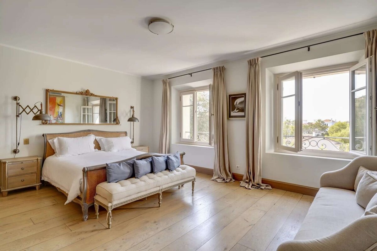 A bright, elegant bedroom with light wood floors, a large bed, a bench, and a sofa. Two open windows reveal a scenic outdoor view.