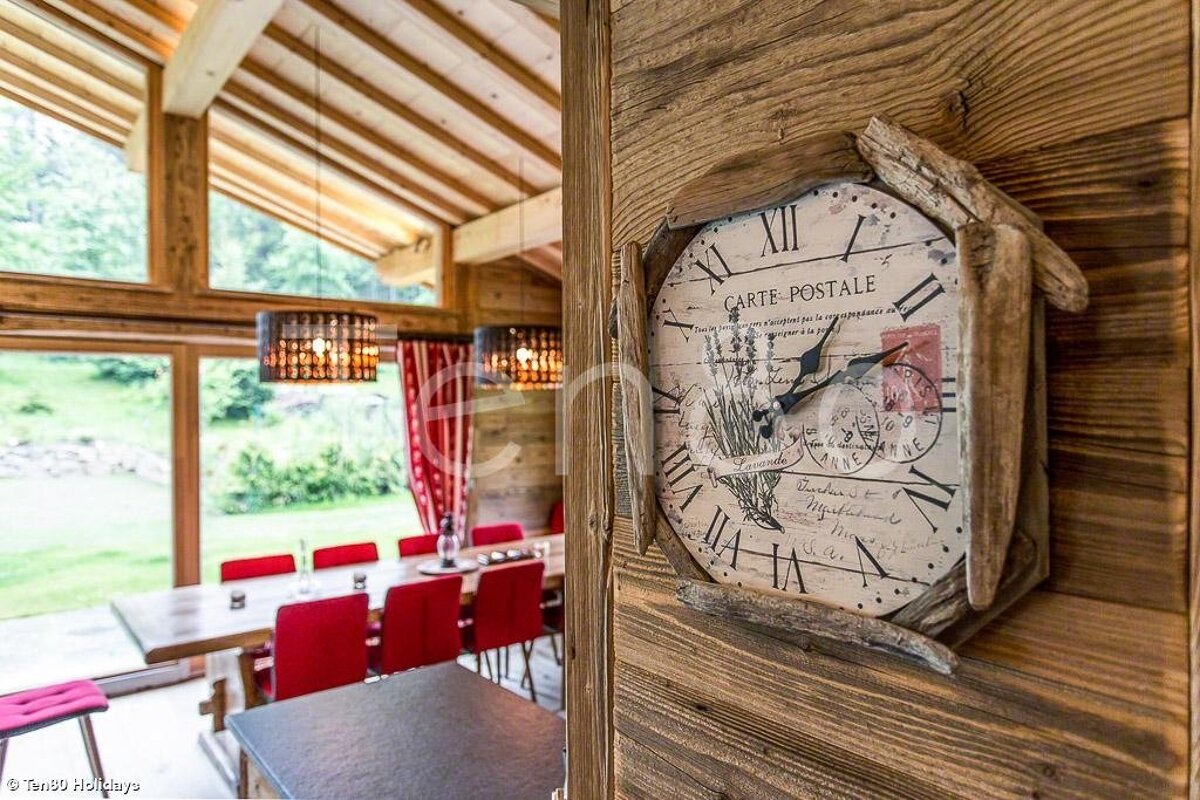 Rustic vintage clock on a wooden wall, with a cozy chalet dining room featuring red chairs in the background.