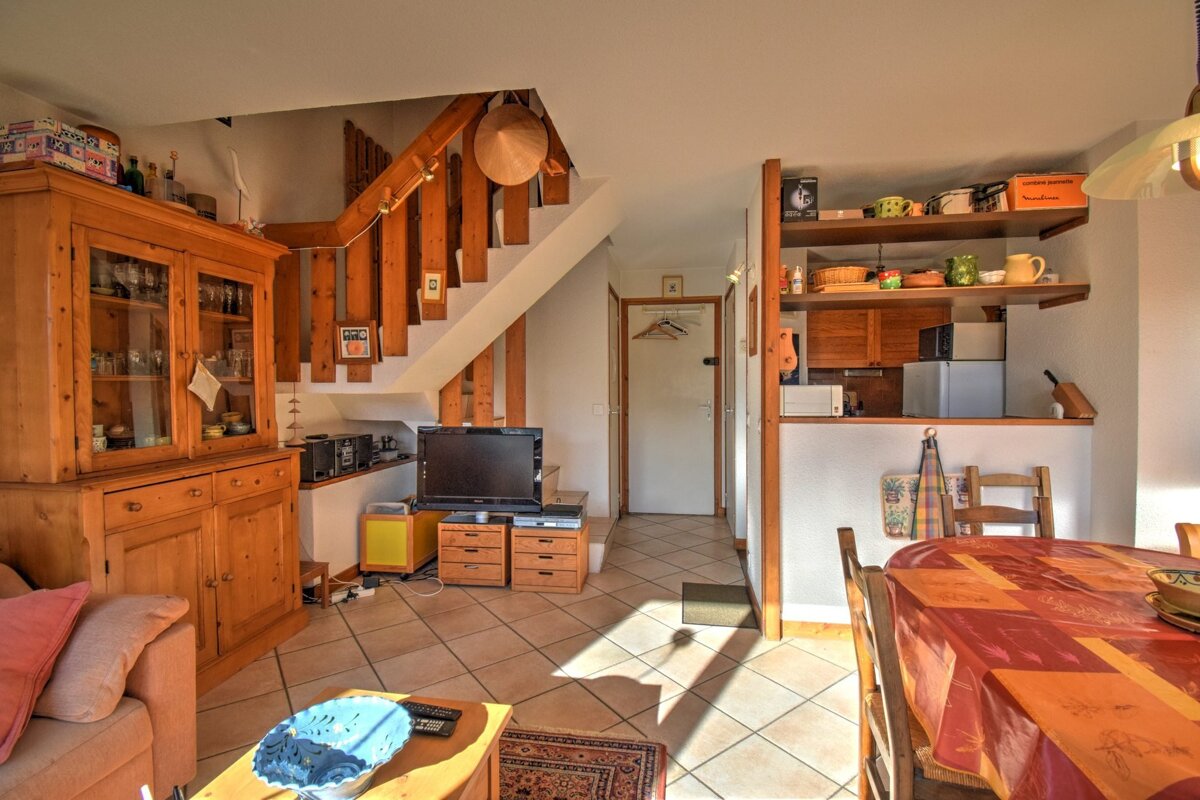 An open-concept living room with a wooden cabinet, staircase, TV area, dining table, and kitchen pass-through. Sunlight hits the tiled floor.