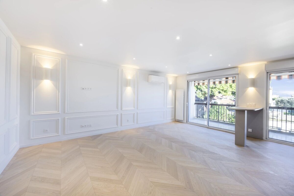 Empty room with white paneled walls, herringbone parquet floors, warm wall sconces, and sliding doors leading to a balcony.