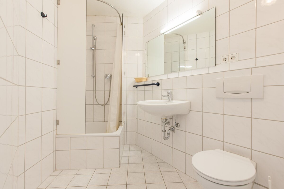 A clean, white-tiled bathroom featuring a bathtub with a shower, a wall-mounted sink, a toilet, and a mirror with an overhead light.