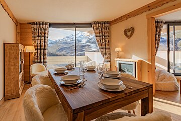 Cozy alpine dining room featuring a wooden table, furry chairs, and a large window showcasing snowy mountain views.