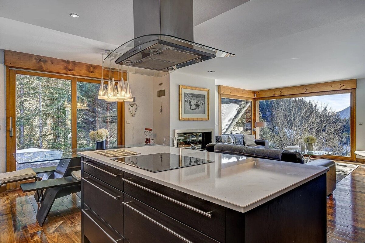 A chic open-plan kitchen with a modern island and living room, boasting large windows overlooking a scenic, snowy forest landscape.