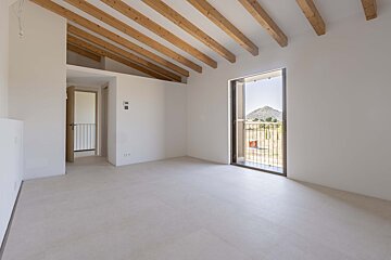 Bright, empty room with white walls, light floor, and exposed wooden ceiling beams. An open balcony door provides a scenic view of a mountain and fields.