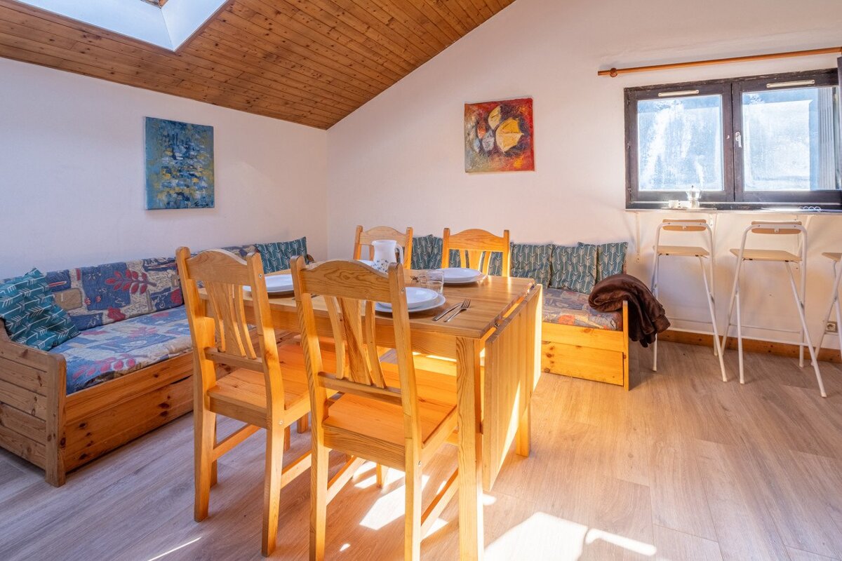 Sunny dining area with a wooden table, chairs, and built-in benches. Features a sloped wood ceiling, skylight, and art, creating a cozy, rustic feel.