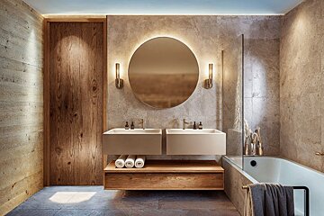 Elegant modern bathroom with a double sink vanity, backlit round mirror, textured stone walls, and warm lighting.