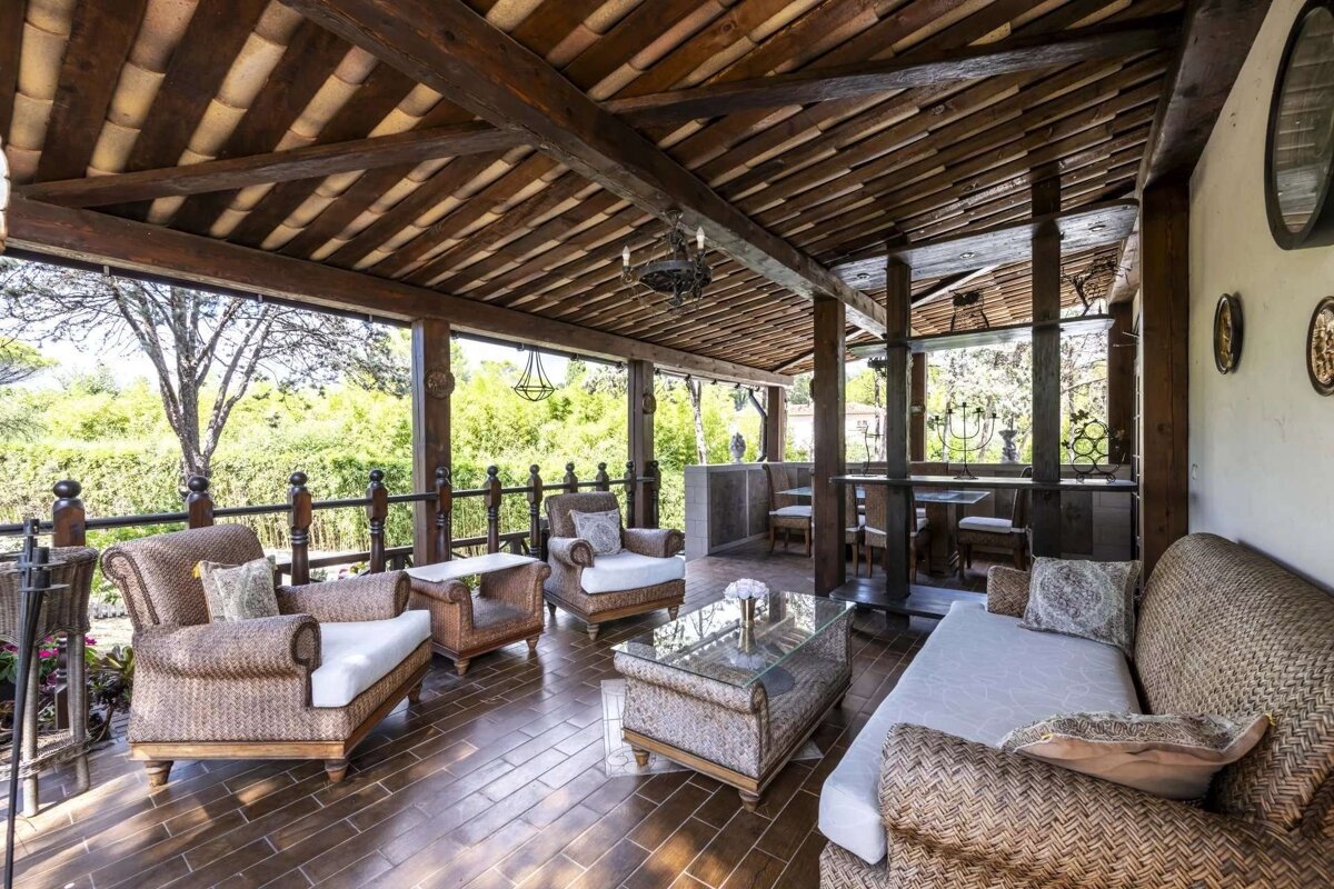 A rustic, covered outdoor patio features woven wicker furniture, a tiled floor, exposed wooden beams, and a view of lush green trees and foliage.