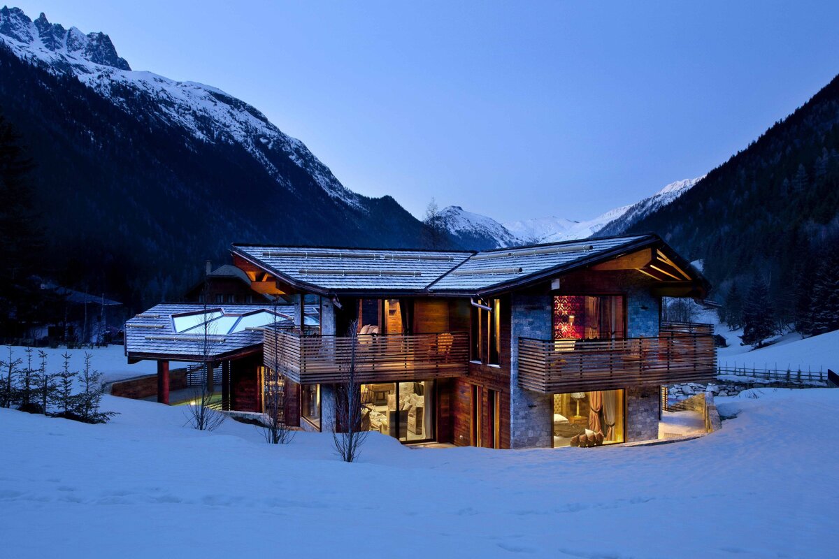 A modern, luxurious wooden chalet glows warmly against a backdrop of dark, snow-capped mountains and pristine snow at twilight.