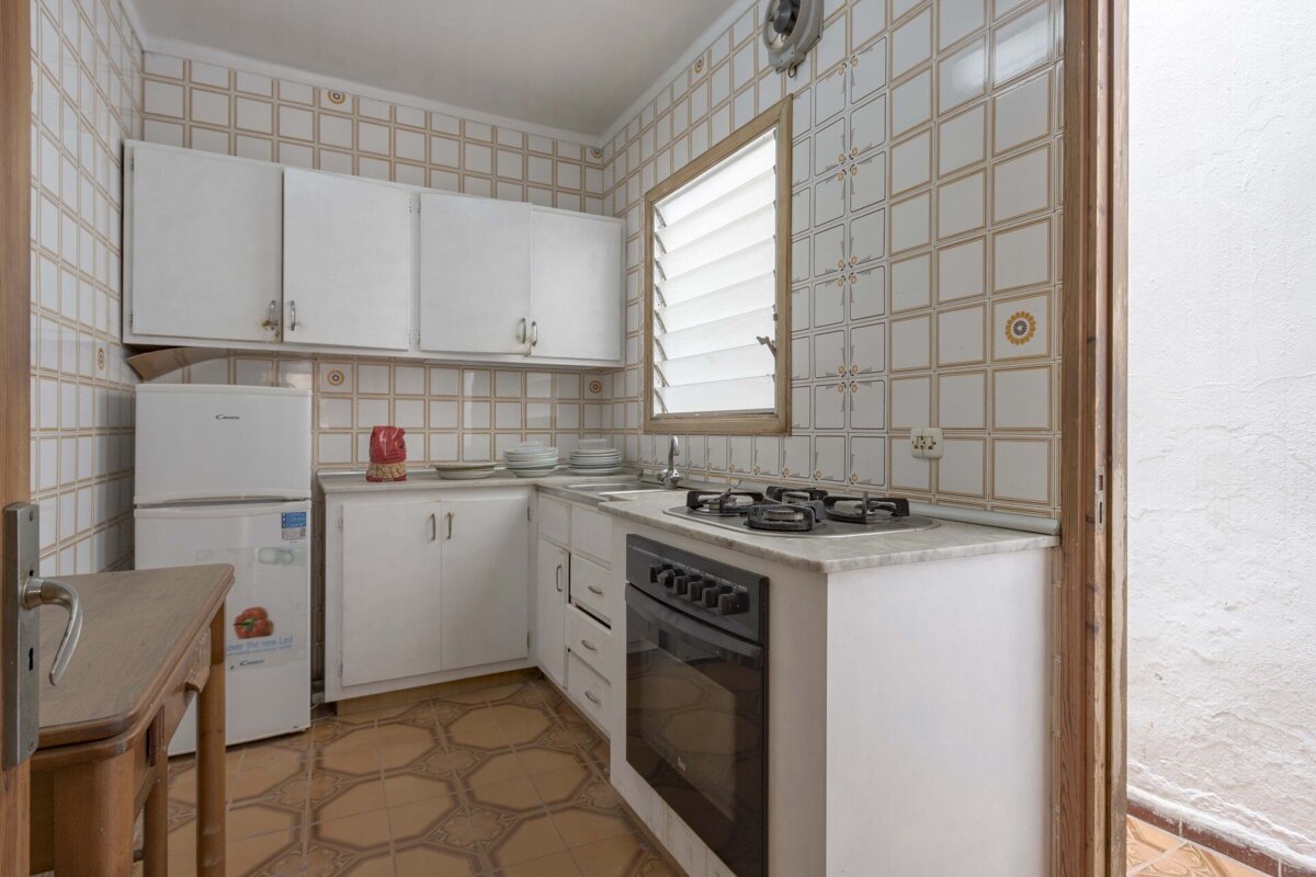 A compact kitchen with white cabinets, a refrigerator, and built-in oven/stove. It features patterned tiled walls and floor, and a louvered window.