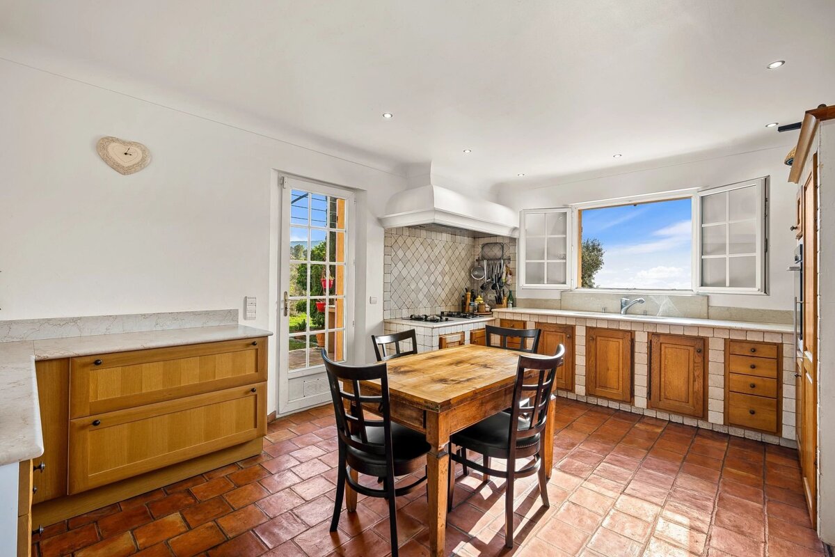 Sunlit rustic kitchen with terracotta floor, wooden dining table, and a large window offering a bright blue sky view.