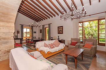 A rustic living/dining room with exposed wood beams, chandeliers, comfortable seating, and a dining table. Large windows offer outdoor views, with a stone archway.