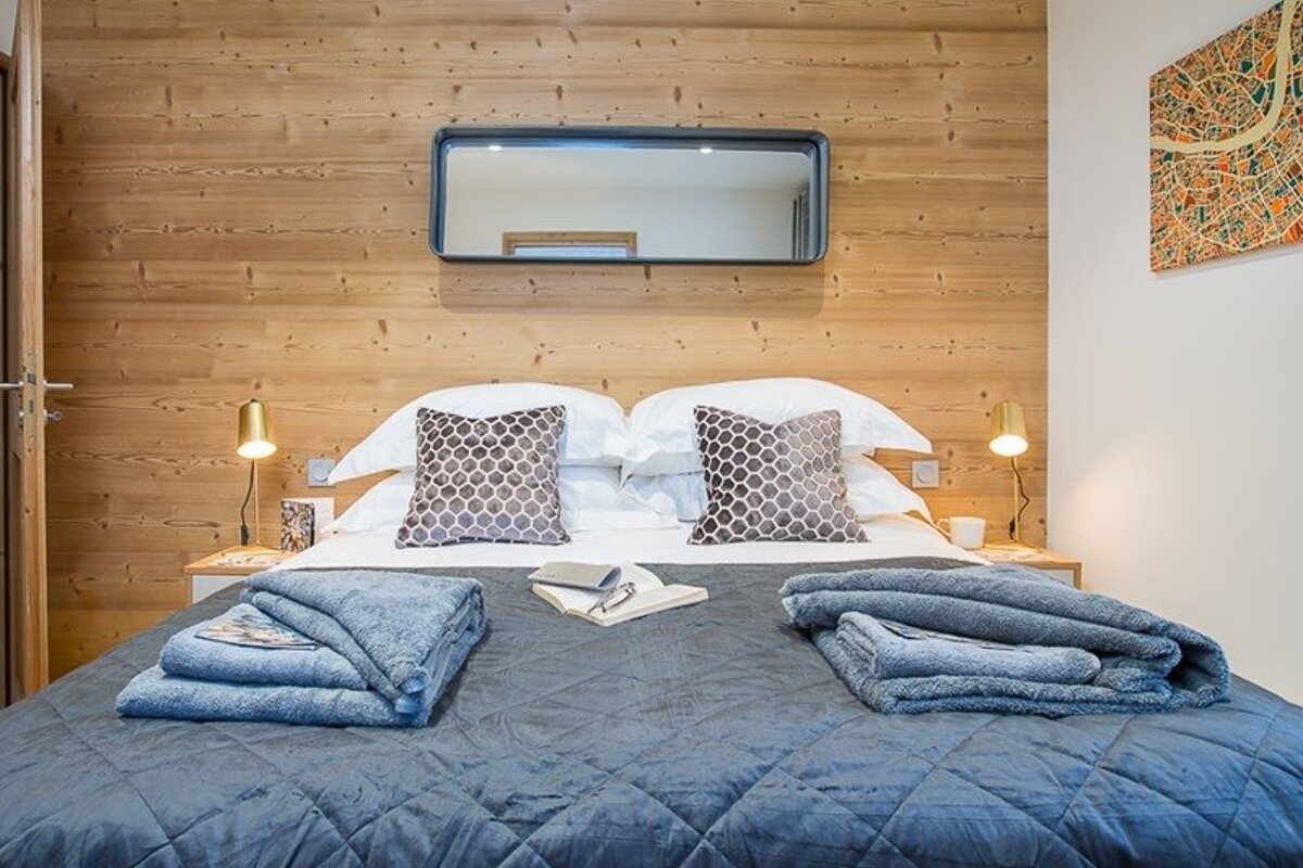 Cozy wood-paneled bedroom with a neatly made bed, blue blankets, patterned pillows, bedside lamps, and a horizontal mirror.