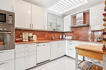 A bright, modern kitchen with glossy white cabinets, red granite countertops, stainless steel appliances, and a skylight. A wooden cart is on the right.
