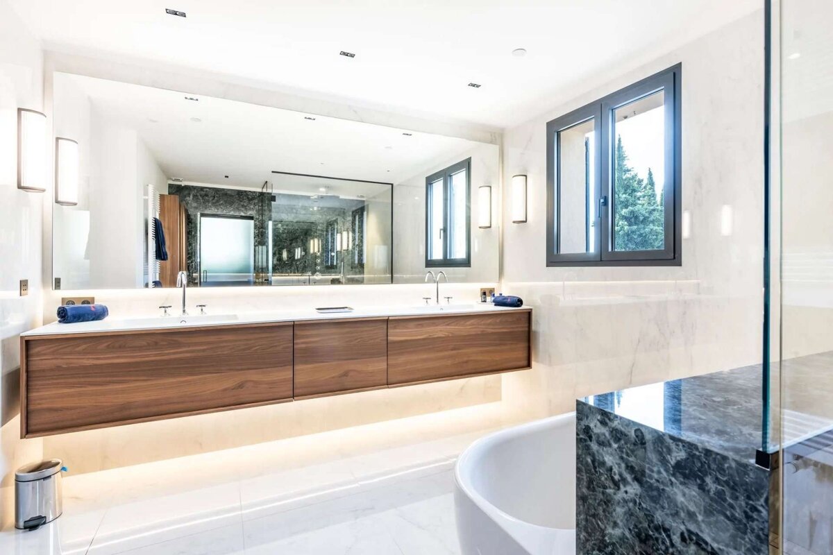 A bright, modern bathroom with a large mirror above a floating wooden double vanity, marble walls, a white tub, and a window looking out to trees.