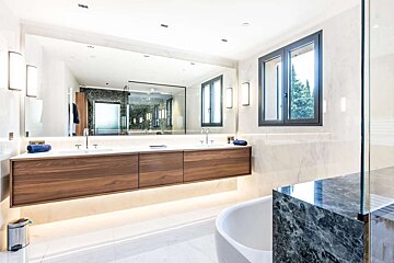 A bright, modern bathroom with a large mirror above a floating wooden double vanity, marble walls, a white tub, and a window looking out to trees.