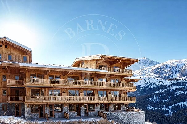 A multi-story wooden alpine chalet with balconies, set against a backdrop of snow-covered mountains and a clear blue sky.