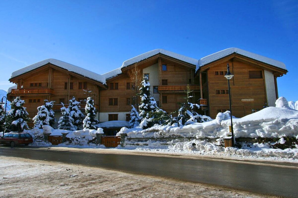 A large, snow-covered wooden chalet building with surrounding snowy trees, set against a clear blue winter sky, beside a partially cleared road.
