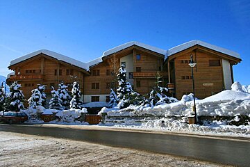 A large, snow-covered wooden chalet building with surrounding snowy trees, set against a clear blue winter sky, beside a partially cleared road.