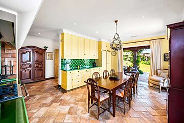 Rustic kitchen-dining room: yellow cabinets, green backsplash, wooden table, terracotta floor, with a garden view.