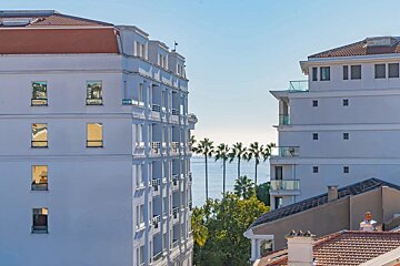 Modern white buildings frame a serene ocean view, dotted with palm trees under a bright blue sky.