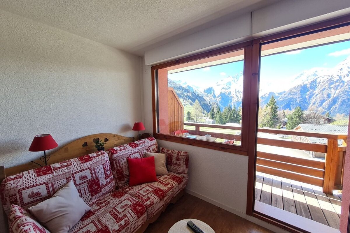 Cozy alpine room featuring a red patterned sofa and large windows opening to a balcony with stunning mountain views.