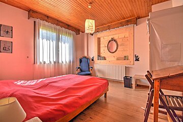 Cozy rustic bedroom featuring a bed with a bright red cover, wooden paneled ceilings, a blue armchair, and a wooden writing desk.