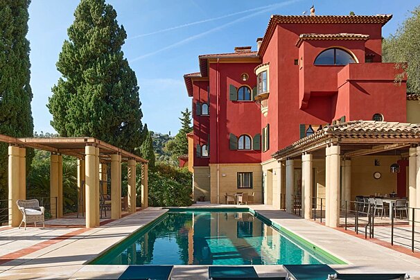 A vibrant red villa with a clear blue swimming pool, tall cypress trees, and covered patios under a sunny sky.