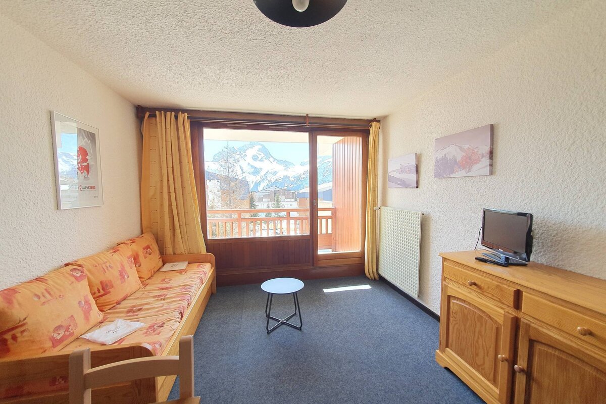 A cozy room features a sofa bed, TV on a wooden cabinet, and a large balcony door revealing a panoramic view of snowy mountains.