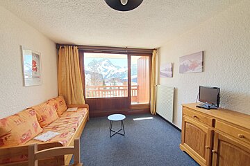 A cozy room features a sofa bed, TV on a wooden cabinet, and a large balcony door revealing a panoramic view of snowy mountains.