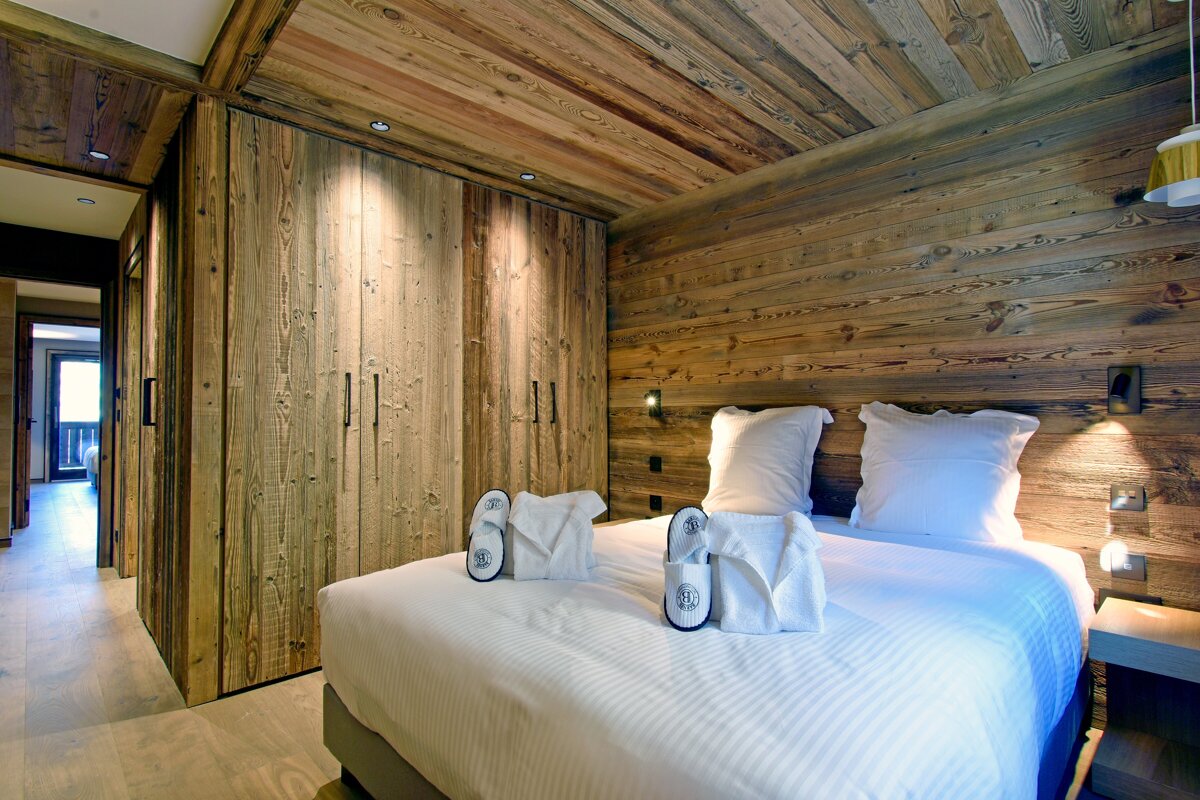 Rustic wooden bedroom with a cozy bed, white linens, robes, slippers, and built-in closet. Warm, inviting mountain lodge style.