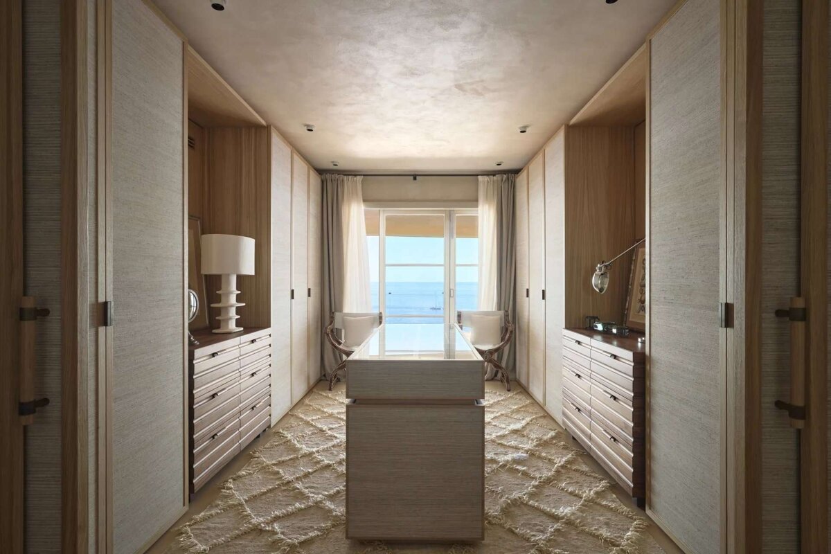 A luxurious walk-in closet in light wood and neutral tones, featuring ample storage, a central island, and a stunning sea view through large windows.