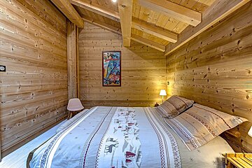 Cozy bedroom with light wood paneling on walls and slanted ceiling. A bed with patterned bedding, two lamps, a chair, and a colorful painting complete the rustic, warm decor.