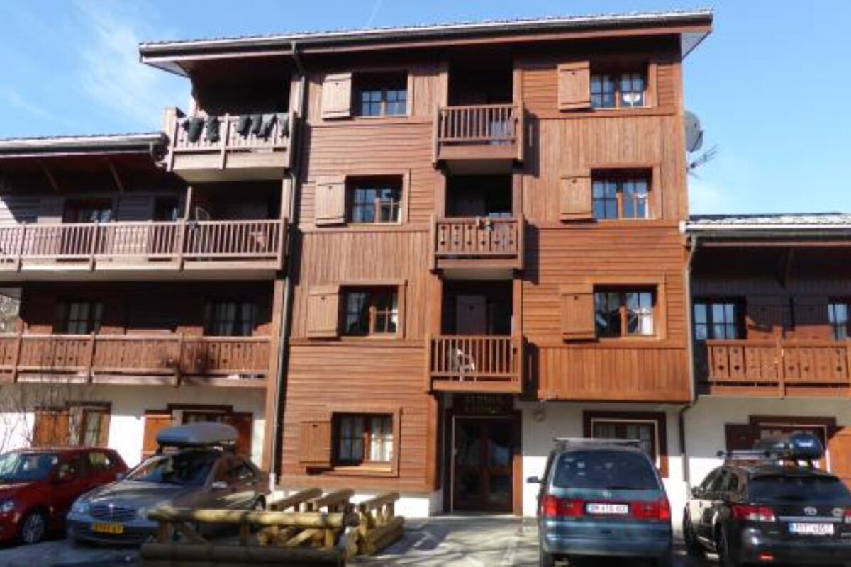 A large, multi-story wooden building with balconies and shutters, cars parked outside on a sunny day.