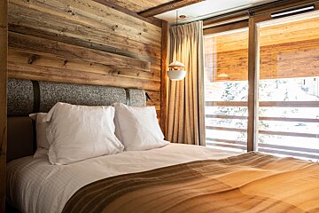 Cozy bedroom with wooden walls, a plush bed with white pillows, and a large window offering a snowy mountain view.